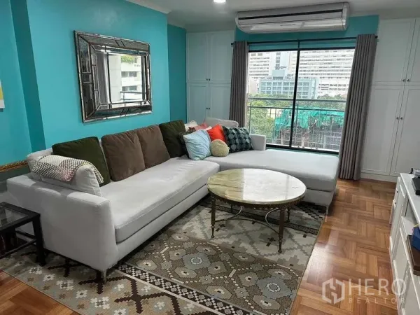 公寓 出售在Bangkok - 明亮客厅，蓝绿色墙面，L形沙发、大理石茶几与城市景观窗。