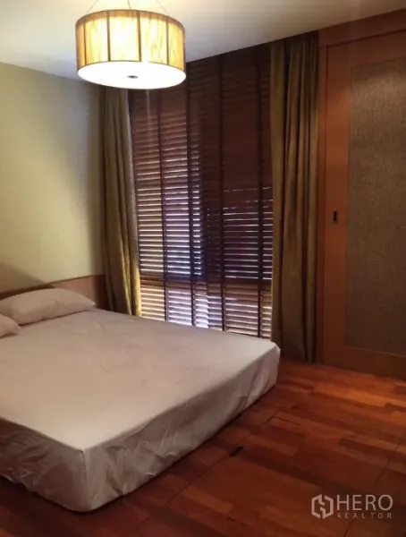 Condominium for rent in Pathum Wan, Bangkok - Cozy bedroom with wooden flooring, window blinds and built-in wardrobe under warm lighting.