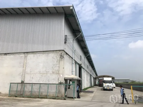 Warehouse for rent in Bang Sao Thong, Samut Prakan - High-clearance metal-clad warehouse exterior with wide concrete yard, loading bay, security gatehouse, and truck access in an industrial zone.