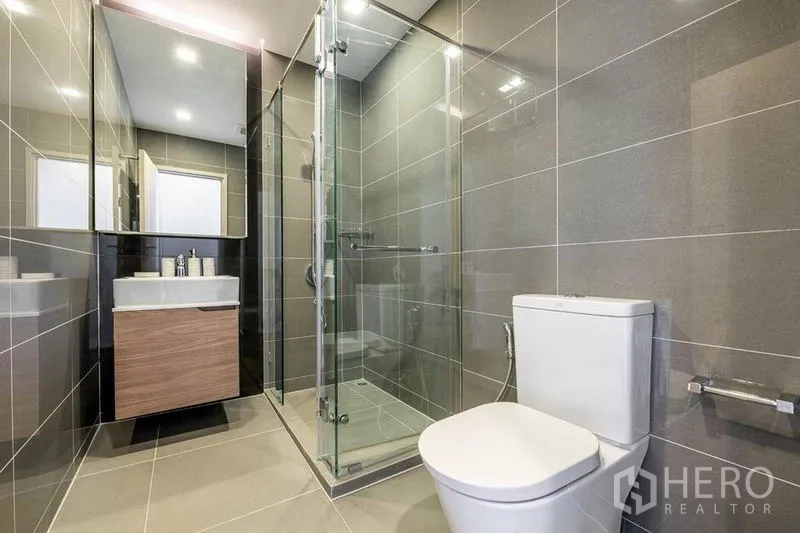 Condominium for rent or sale in Watthana, Bangkok - Sleek bathroom featuring a glass-enclosed shower, gray tiles, vanity and toilet.