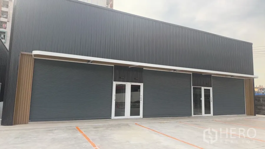 货栈 出租在Prawet, Bangkok - 现代仓库外立面，深色金属板、玻璃门、卷闸门与标线停车位。
