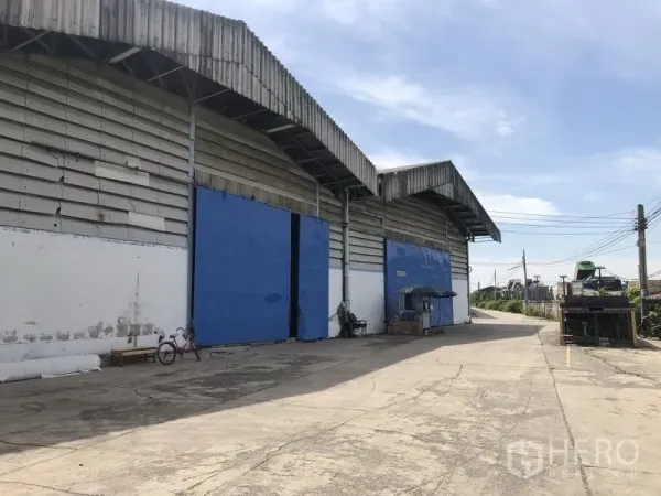 Warehouse for rent in Bang Phli, Samut Prakan - Row of warehouses with wide driveway and multiple blue loading doors for easy truck access.