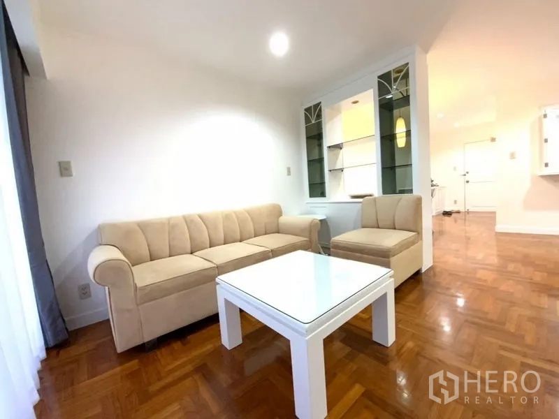公寓 出租在Watthana, Bangkok - 温馨起居角，米色转角沙发与白色茶几，旁有展示架。