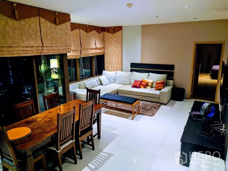 Condominium for rent - Open-plan living room with L-shaped sofa, wooden dining set, and large windows with Roman blinds at night.