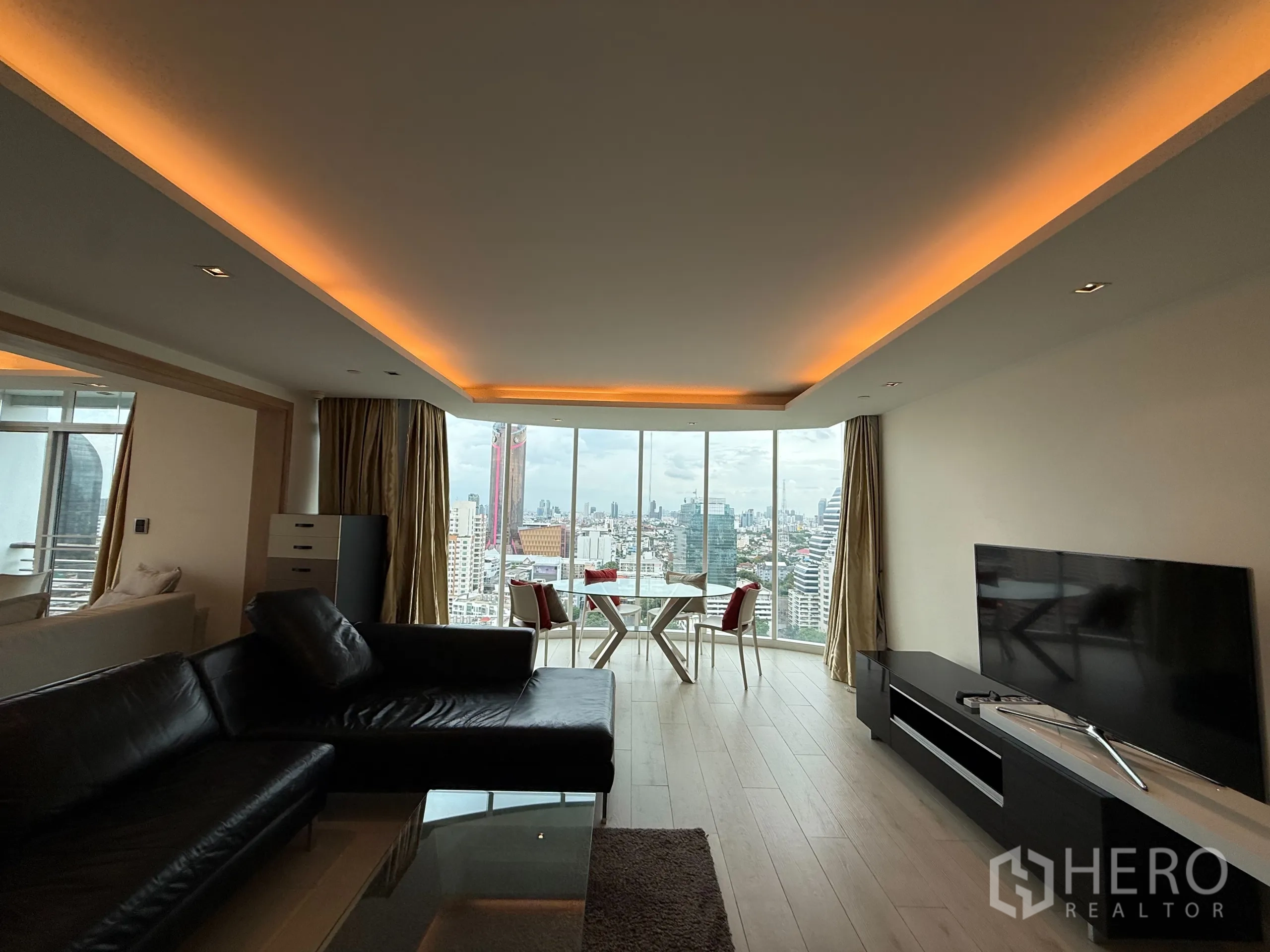 Condominium for rent in Phaya Thai, Bangkok - Long living room with cove lighting, sectional sofa and TV facing expansive city views through full‑height glazing.