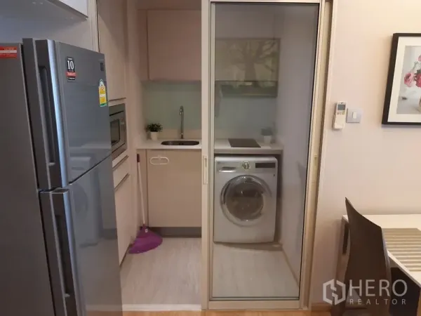 公寓 出租在Watthana, Bangkok - 推拉玻璃门分隔的小厨房，带灶台、水槽和前置洗衣机。