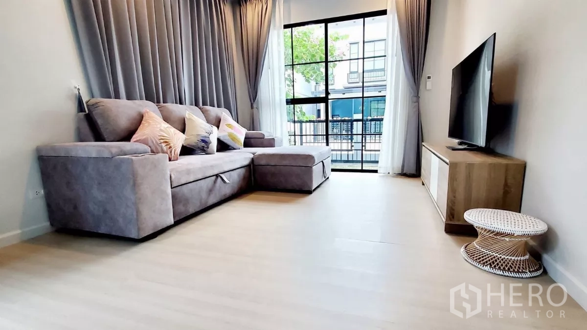Detached House for rent in Lat Krabang, Bangkok - Cozy living room with grey sectional sofa facing a flat-screen TV and a floor-to-ceiling sliding glass door.