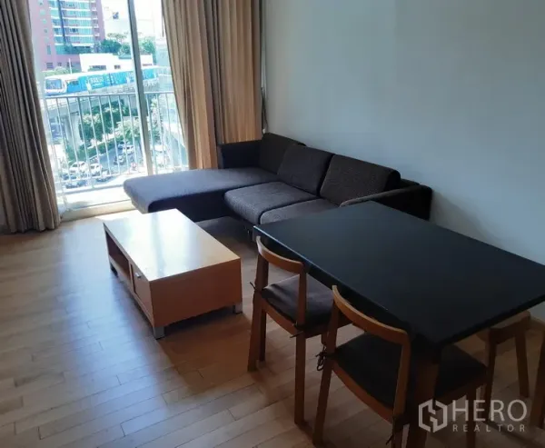 Condominium for rent in Khlong Toei, Bangkok - Bright living and dining area with sectional sofa, dining table, wood floors and a balcony with city view.