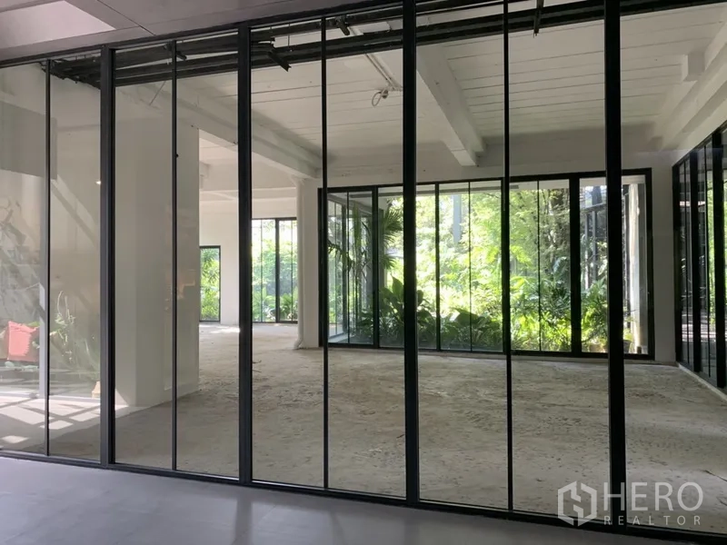 Office Space for rent in Khlong Toei, Bangkok - Sunlit office behind a glass façade with garden backdrop and exposed ceiling beams.