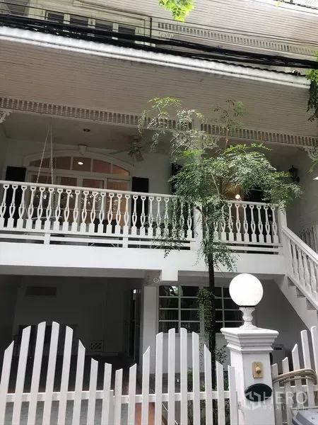 Townhouse for rent in Mueang Samut Prakan, Samut Prakan - White two-storey townhouse exterior with balcony, decorative railings, gated entry and leafy tree.