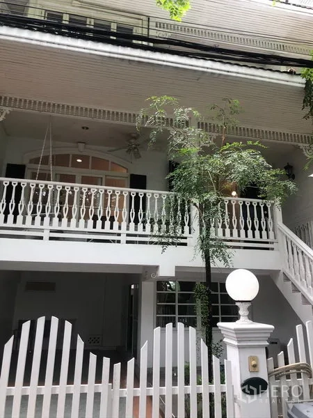 Townhouse for rent in Mueang Samut Prakan, Samut Prakan - White two-storey townhouse exterior with balcony, decorative railings, gated entry and leafy tree.