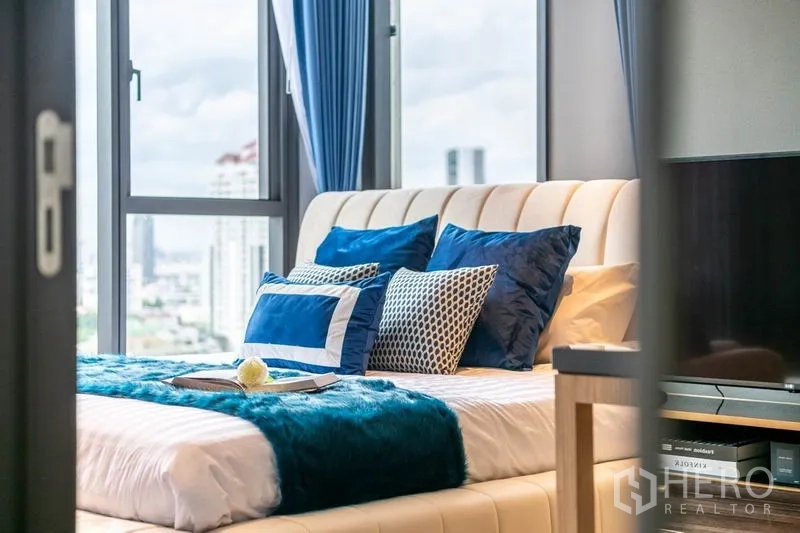 Condominium for rent or sale in Khlong Toei, Bangkok - Close-up of bed with teal throw and pillows next to large windows.