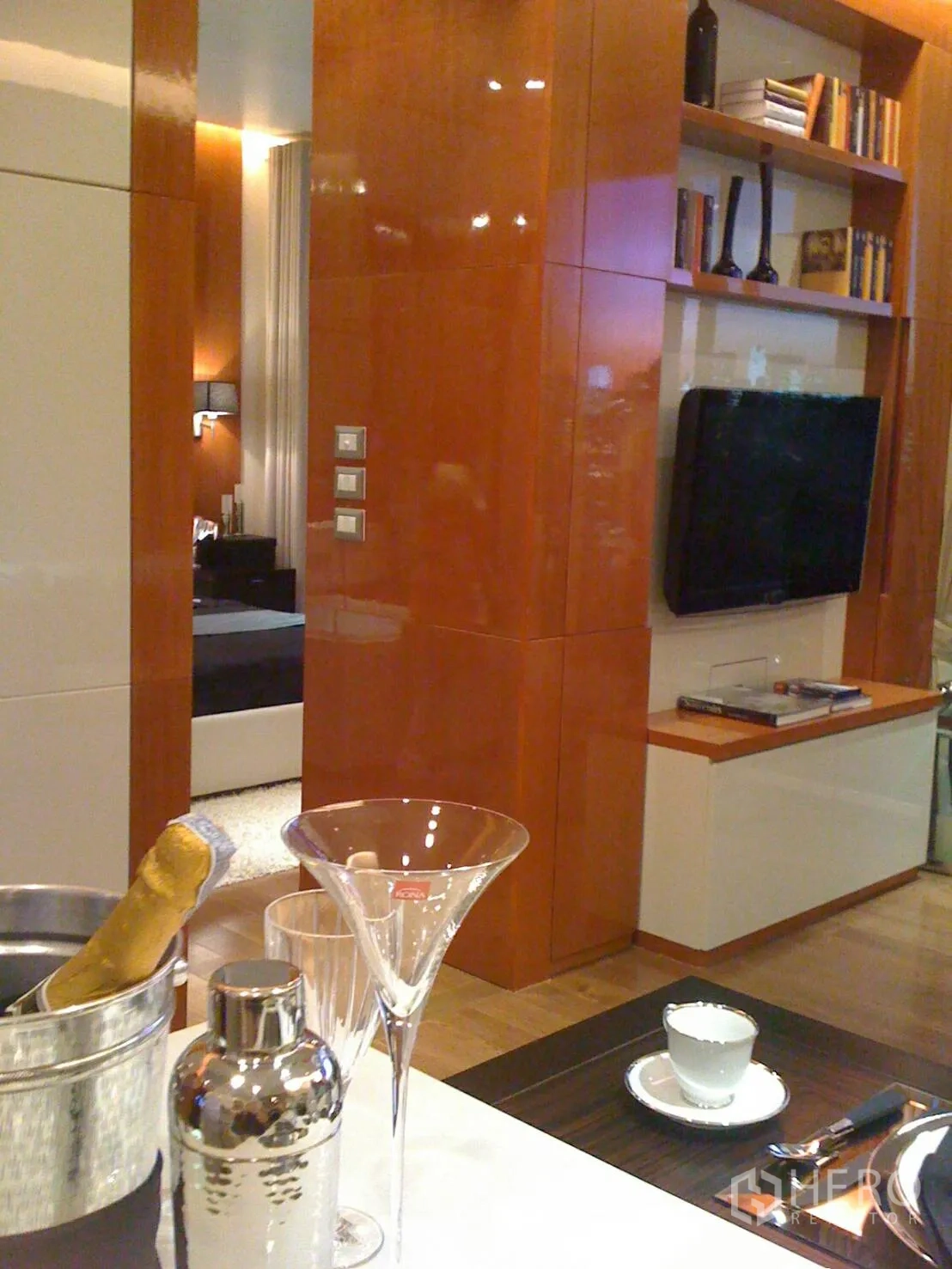Condominium for rent or sale in Khlong Toei, Bangkok - Open-plan living with glossy wood panels, built-in shelving, wall-mounted TV, and breakfast bar with glassware.