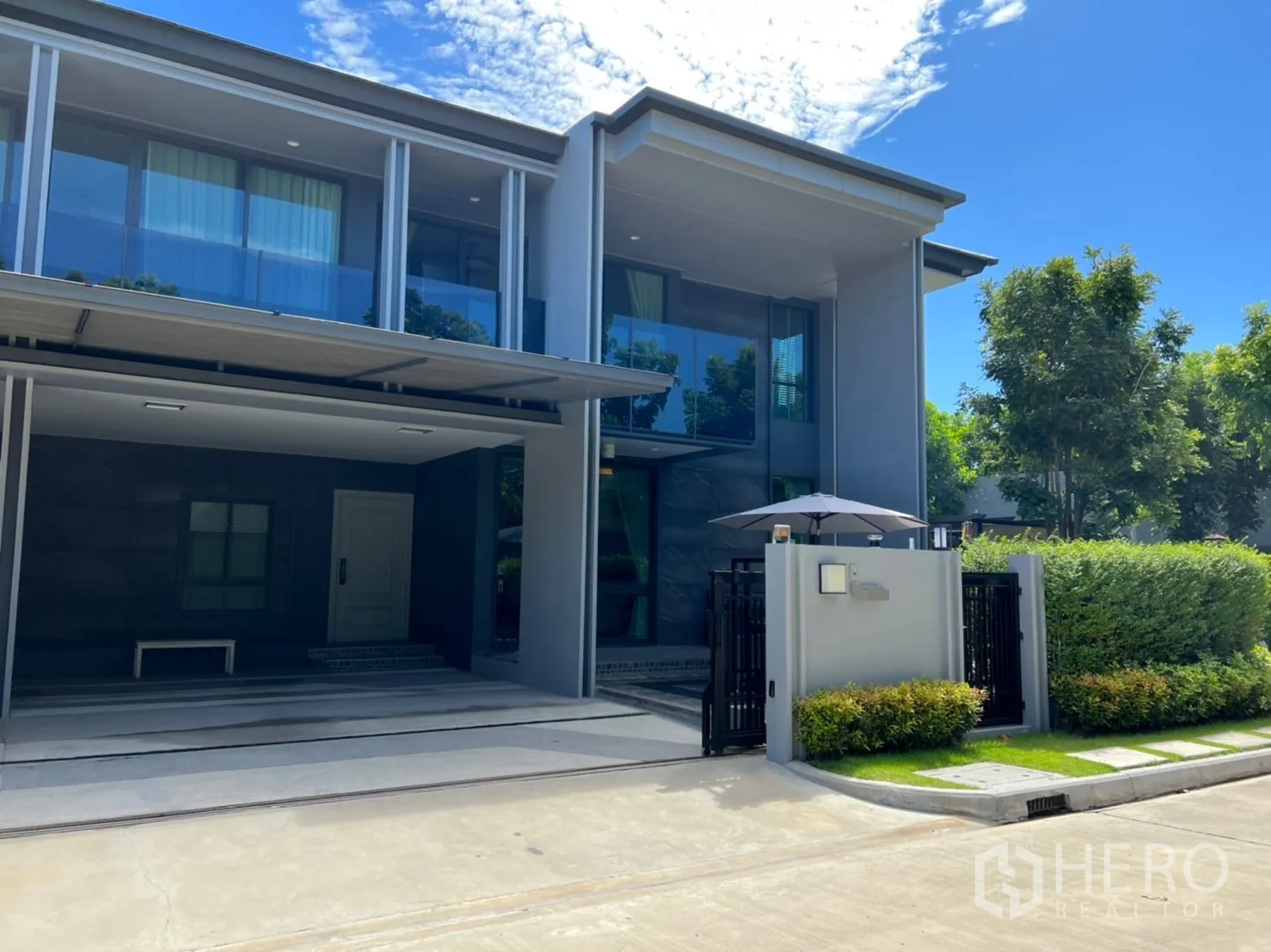 Detached House for rent or sale in Bang Kapi, Bangkok - Modern two-storey detached house exterior in Bang Kapi, Bangkok with glass balcony, covered carport and landscaped hedge under a blue sky.