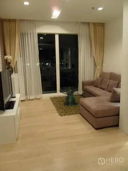 Condominium for rent in Khlong Toei, Bangkok - Living area with L-shaped sofa, glass coffee table and balcony doors showing city night view.