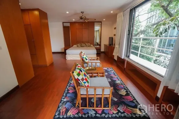 Apartment for rent in Watthana, Bangkok - Large master bedroom with sitting area, floor-to-ceiling window and warm wood built-ins overlooking greenery.