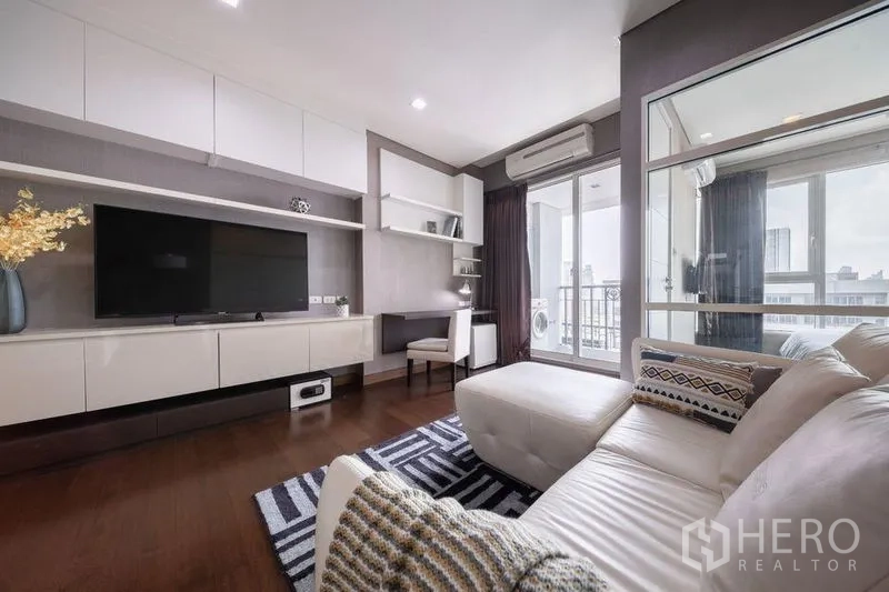 Condominium for rent or sale in Watthana, Bangkok - Modern living room with L-shaped sofa, shelving, TV console and door to balcony.