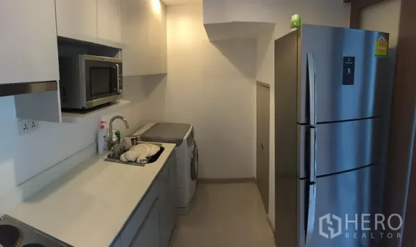 Condominium for rent in Phra Khanong, Bangkok - Modern galley kitchen with white cabinets, stainless refrigerator, microwave, and washer.