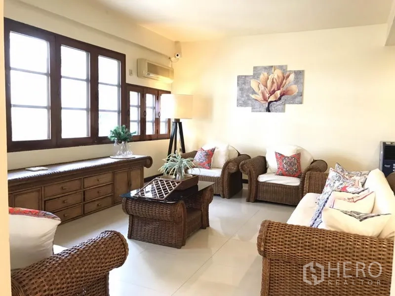 Detached House for rent in Phra Khanong, Bangkok - Airy lounge with rattan sofas, artwork, and rows of wooden-framed windows.