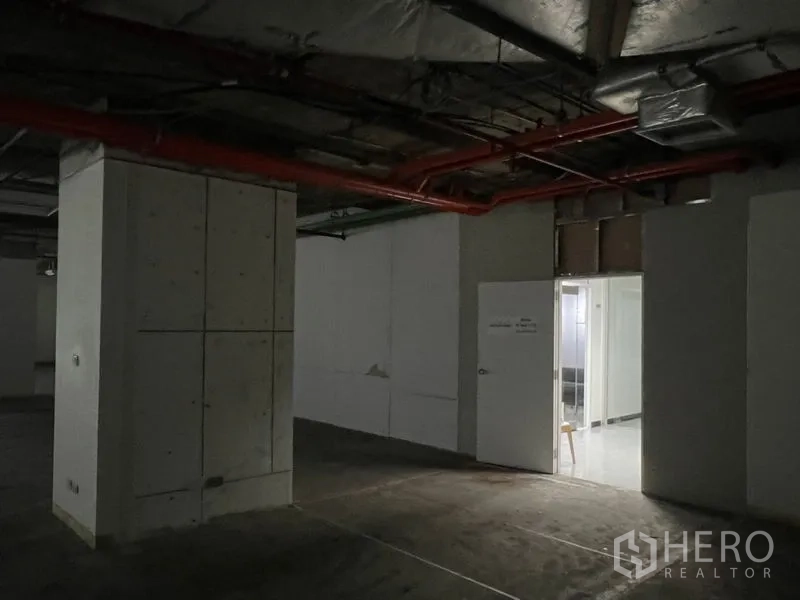 办公空间 出租 - 昏暗的室内走廊，结构立柱与红色消防管，通向更明亮的区域。