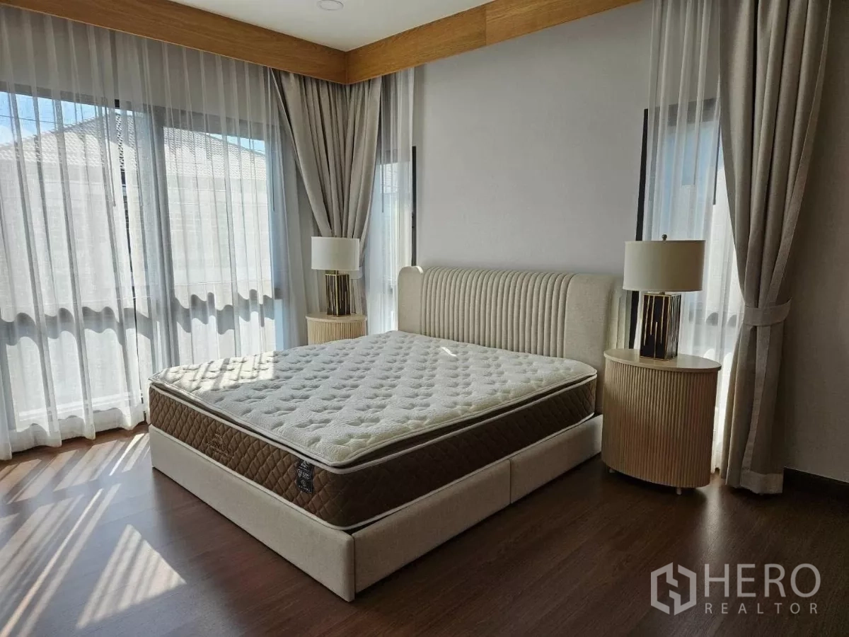Detached House for rent or sale in Bang Phli, Samut Prakan - Sunlit bedroom with large corner windows, sheer curtains, upholstered bedframe, and wood flooring.