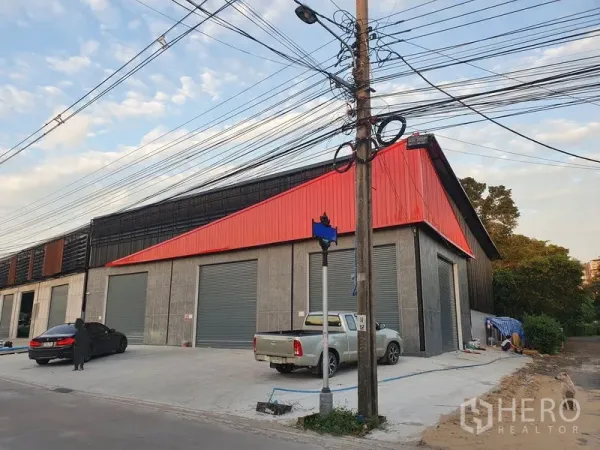 Warehouse for rent in Pak Kret, Nonthaburi - Corner view of a modern warehouse with red cladding, multiple roller shutters and a wide concrete loading apron.
