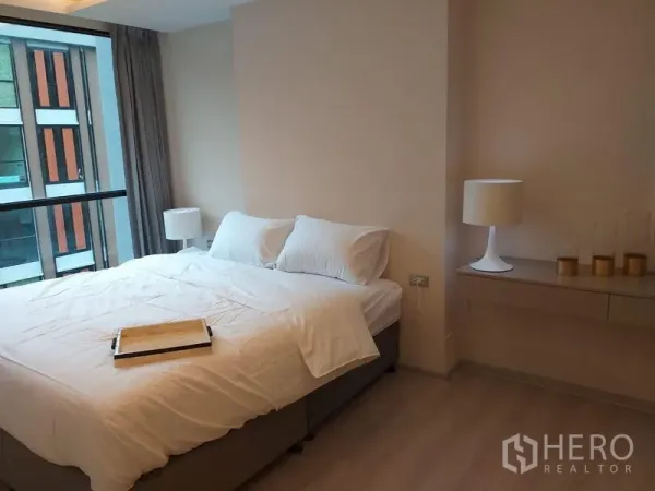 Condominium for rent in Khlong Toei, Bangkok - Primary bedroom with large bed, bedside lamps, and a wide window bringing in natural light.