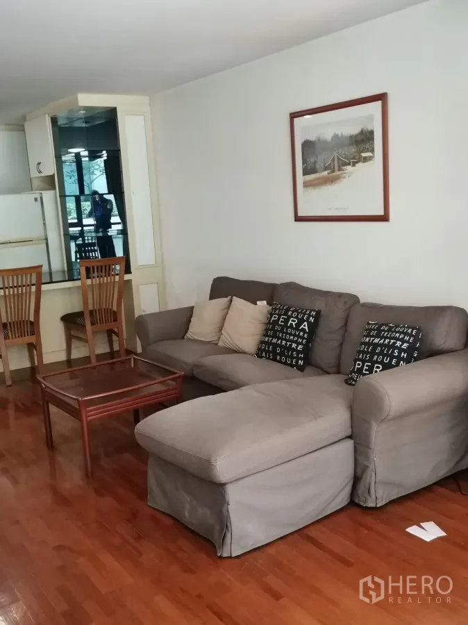 公寓 出租在Pathum Wan, Bangkok - 开放式客厅，灰色转角沙发与茶几，木地板，旁有镜面柜增添采光。
