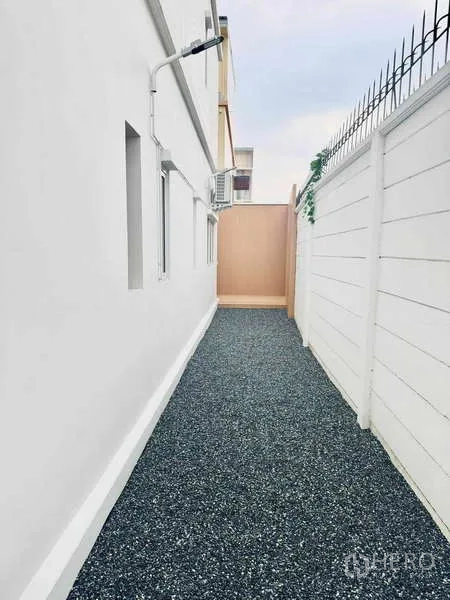 Townhouse for rent in Suan Luang, Bangkok - Side yard walkway with gravel surface, a high white privacy wall, and exterior air-conditioning units.