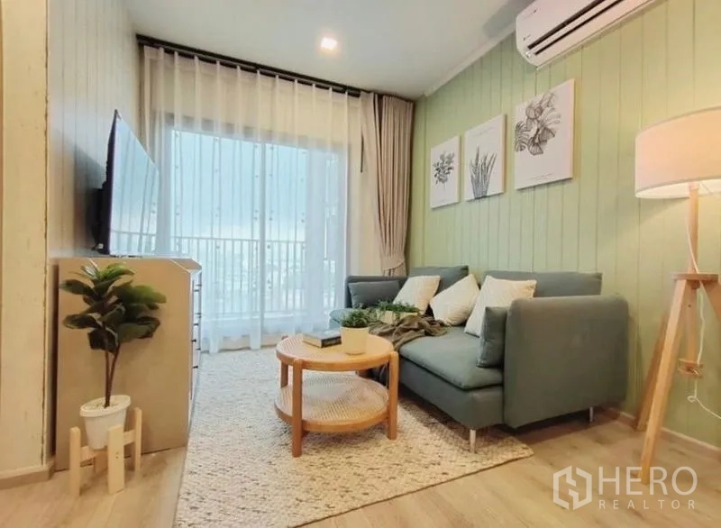 คอนโด ให้เช่าหรือขาย - ห้องนั่งเล่นเปิดออกสู่ระเบียง ม่านโปร่ง ทีวีติดผนัง และผนังสีเขียวเด่น