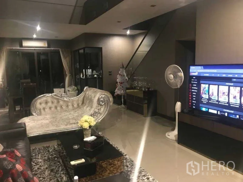 Townhouse for rent - Spacious living area at night with large TV, dining nook, and staircase with glass railing.