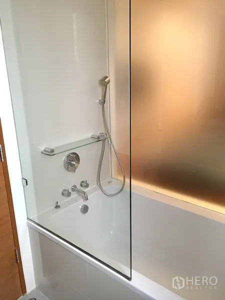 Condominium for rent or sale in Khlong Toei, Bangkok - White bathtub with handheld shower and glass screen in a contemporary bathroom.