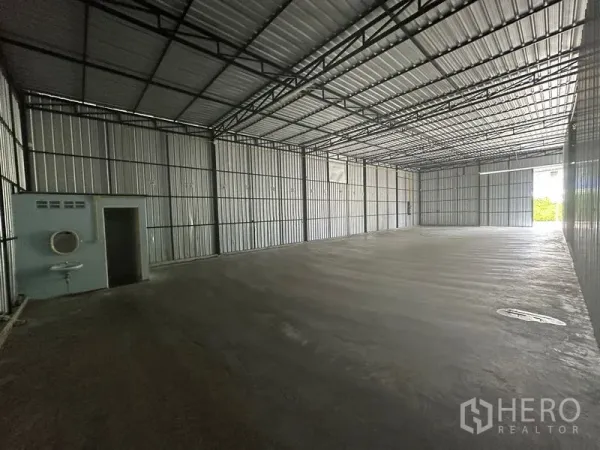 Warehouse for rent in Wang Thonglang, Bangkok - Spacious empty warehouse interior with high steel truss roof, concrete floor, and a small restroom and sink along one wall.