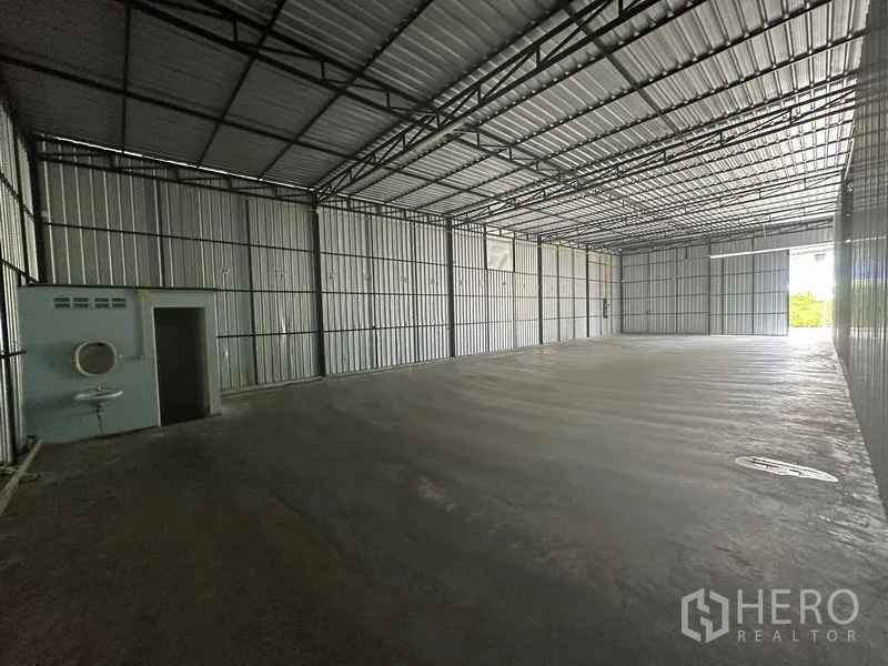 Warehouse for rent in Wang Thonglang, Bangkok - Spacious empty warehouse interior with high steel truss roof, concrete floor, and a small restroom and sink along one wall.