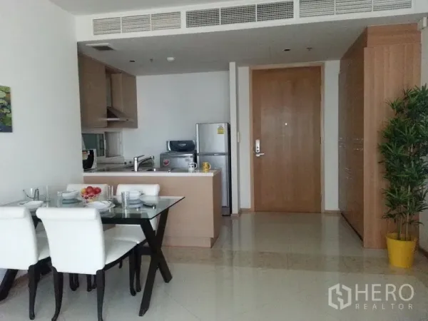 Condominium for rent in Sathon, Bangkok - Open-plan kitchen with light wood cabinets, island, and a glass dining table by the entry in a Sathon condo.