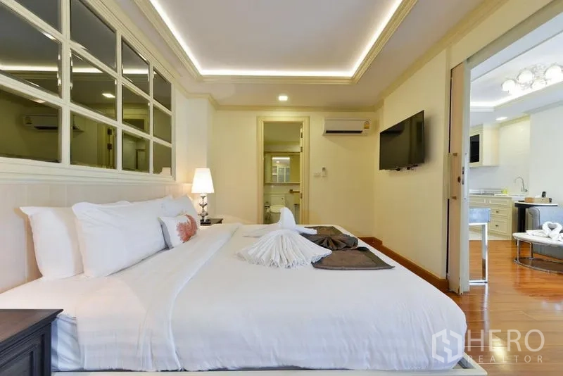 Apartment for rent in Khlong Toei, Bangkok - Cozy bedroom with king bed, mirrored wall, LED cove lighting, and doorway to bathroom.