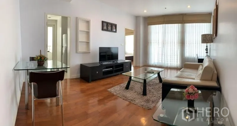 Condominium for rent in Khlong Toei, Bangkok - Open‑plan living area with TV console, sofa set, glass tables, rug, and full‑height windows.