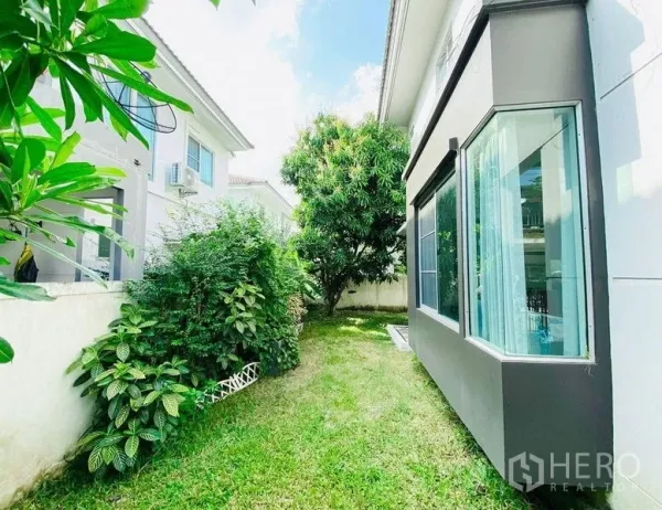 独立式住宅 出租 - 曼谷Prawet区现代独栋别墅侧院，草坪、灌木与成树在阳光下。