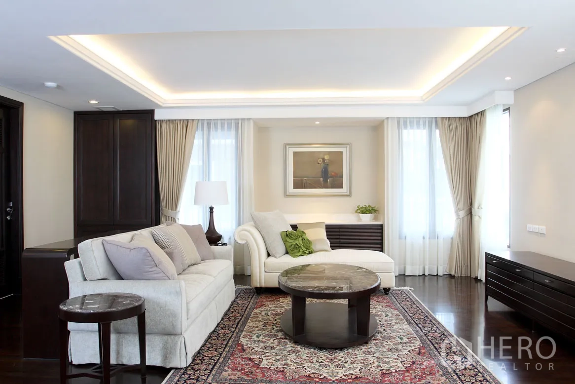 Detached House for rent in Khlong Toei, Bangkok - Light-filled living room with coffered ceiling, sofas, patterned rug and wide windows with drapes.