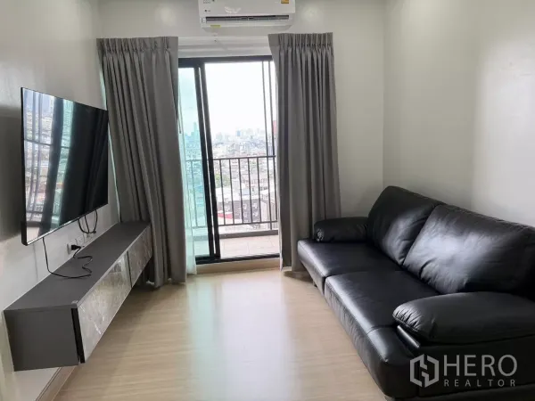 公寓 出租在Khlong Toei, Bangkok - 客厅配黑色皮沙发、壁挂电视与通往阳台的玻璃门，城市景观。