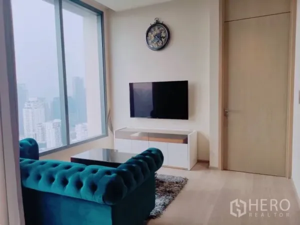Condominium for rent or sale in Watthana, Bangkok - Cozy living room with teal tufted sofa, wall-mounted TV and a large window overlooking the city.