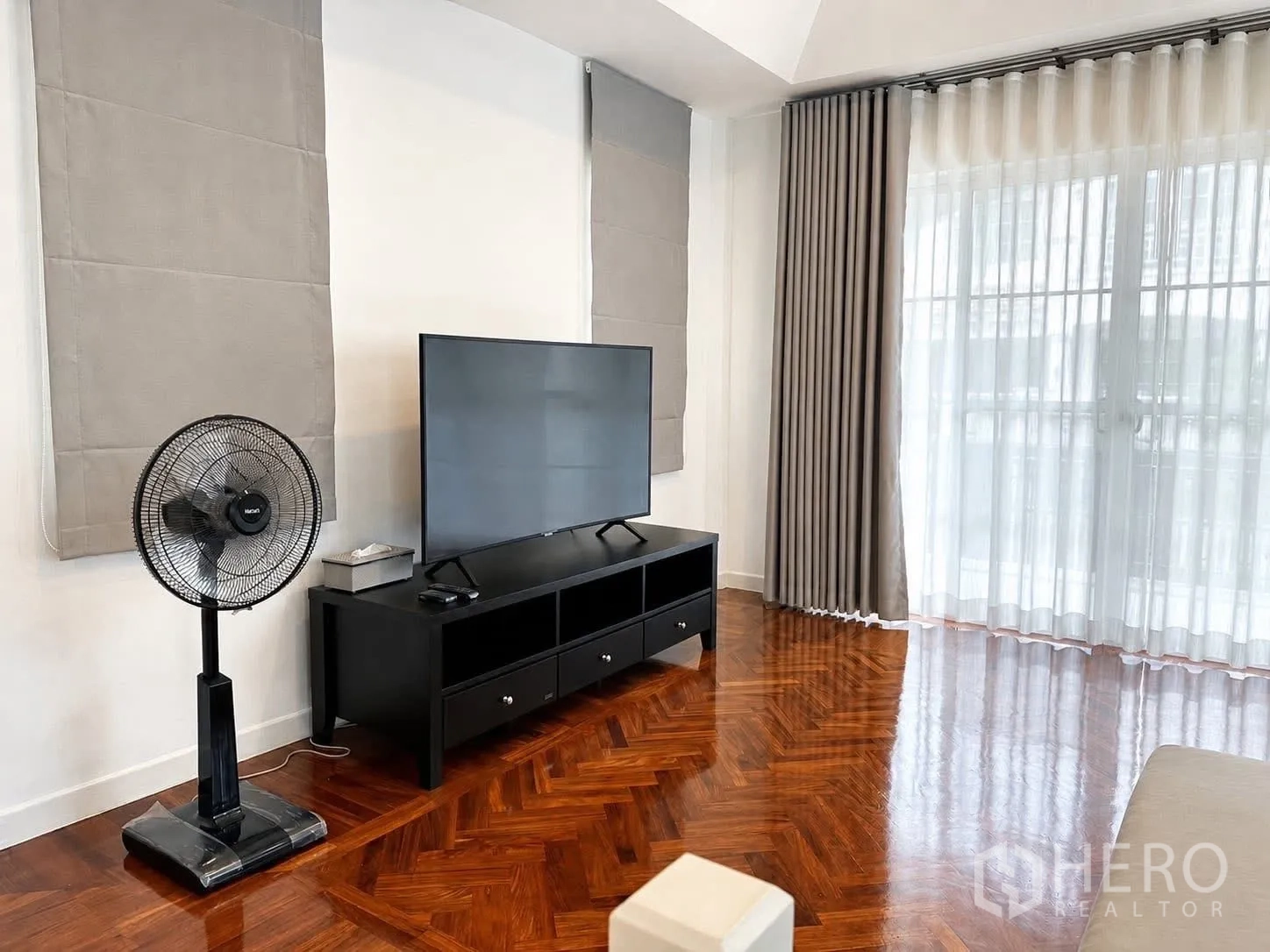 独栋房屋 for rent in Prawet, Bangkok - 影音区有电视柜、立式风扇与人字拼木地板，旁为玻璃门。