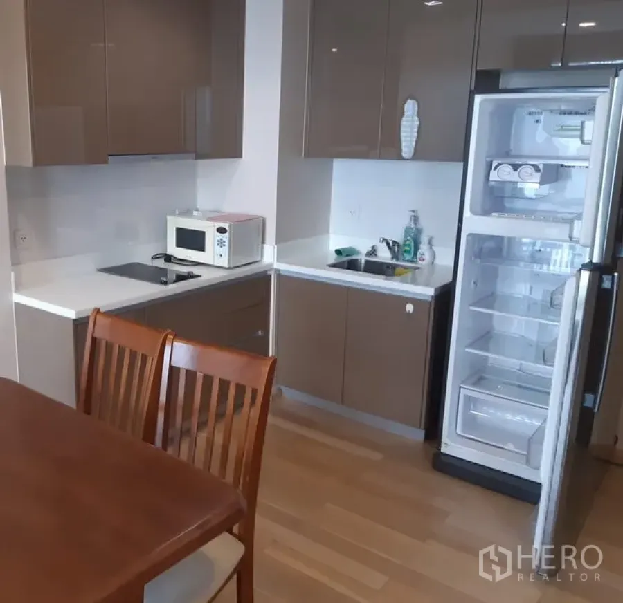 Condominium for rent in Khlong Toei, Bangkok - Modern compact kitchen with glossy brown cabinets, white countertops, built-in hob, microwave, sink, and open refrigerator.