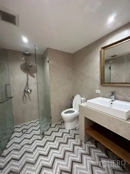 Condominium for rent in Khlong Toei, Bangkok - Modern bathroom with glass-enclosed shower, toilet and vanity with square sink on chevron tile flooring.