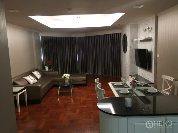 Condominium for rent in Khlong Toei, Bangkok - Open-plan living and dining with L-shaped sofa, wall-mounted TV, breakfast bar, and recessed ceiling lighting.