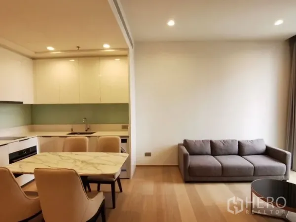 Condominium for rent in Bang Rak, Bangkok - Open-plan living area with gray sofa, stone-top dining table and a white L-shaped kitchen with built-in appliances and wood flooring.