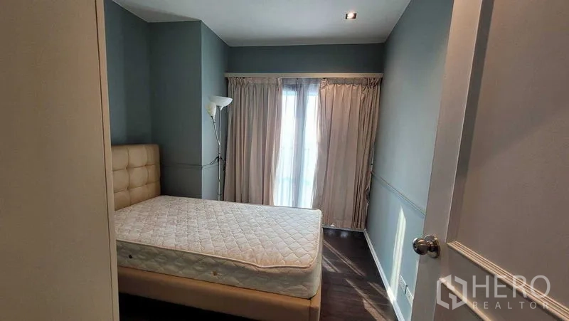 Condominium for rent in Khlong Toei, Bangkok - Cozy bedroom with upholstered bed, dark wood floor and floor-to-ceiling curtains opening to a balcony.
