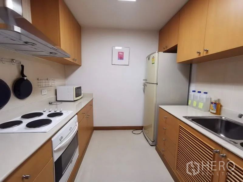 Condominium for rent in Pathum Wan, Bangkok - Galley kitchen with electric stove, oven, range hood, microwave, fridge and light wood cabinets.