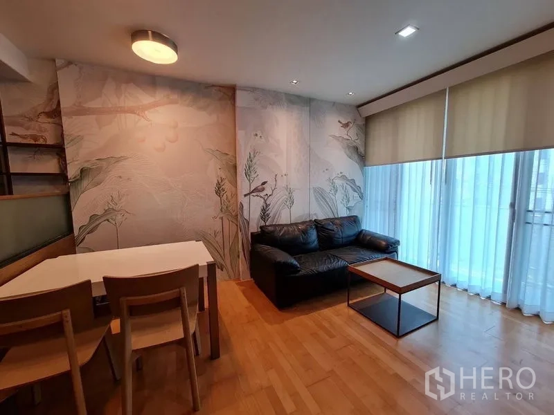 Condominium for sale in Khlong Toei, Bangkok - Bright living room with leather sofa, mural wallpaper and floor-to-ceiling windows with blinds.
