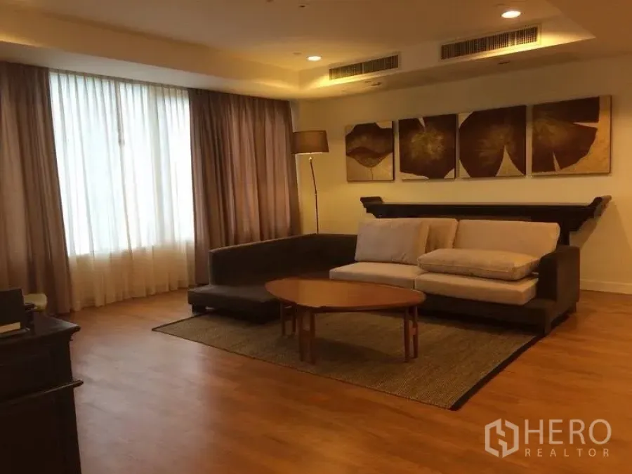Condominium for rent in Watthana, Bangkok - Warm living room with L-shaped sofa, coffee table, floor lamp, wall art and large window.